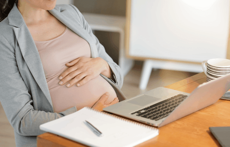 Een zwangere vrouw zit aan een houten tafel met een laptop en een schrift.