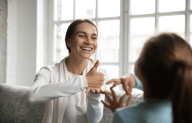 Een lachende vrouw met in een witte trui leert een meisje gebaren tijdens een behandeling logopedie.